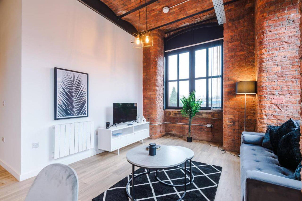 Meadow Mill Stockport living room space with a plant in the exposed brickwork window, tv on a stand on. white wall and coffee table in the centre atop a black and white rug